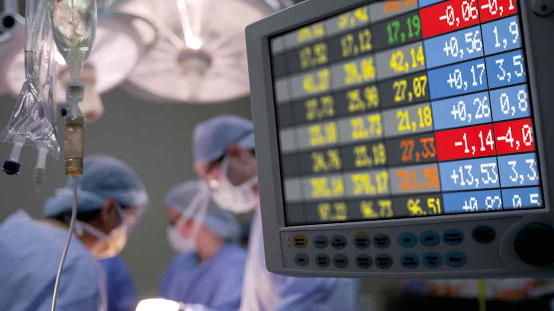 Ärzte am OP-Tisch, im Vordergrund ein Monitor mit Wirtschaftsdaten