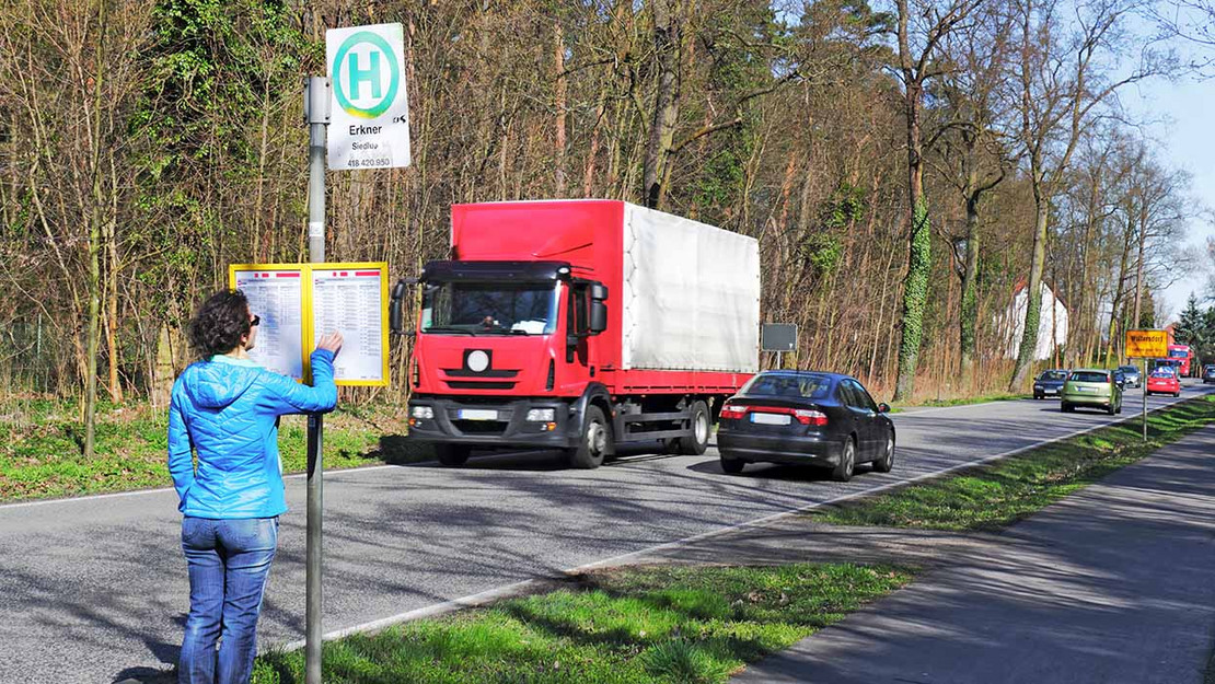 Eine Person steht an einer Bushaltestelle an einer stark befahrenen Landstraße und liest den Fahrplan. 