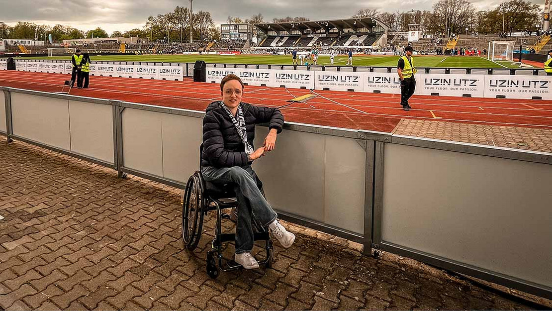 Junger Mann im Rollstuhl sitzt an einer Balustrade. Im Hintergrund eine Laufbahn und ein Fußballplatz. 