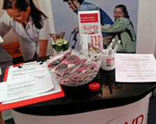 Messe-Counter mit Faltblättern, Stiften, roten Sonnenbrillen und Pfefferminzdosen des SoVD. Poster im Hintergrund zeigen eine Pflegefachkraft und ein junges Paar mit Behinderung auf einem Fahrrad.