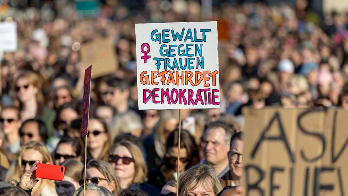 Große Demonstration. Mittig wird ein Plakat hochgehalten mit der Aufschrift: Gewalt gegen Frauen gefährdet Demokratie.