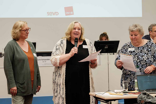 Drei Frauen stehen nebeneinander; die mittlere hält ein Papier in der Hand und spricht ins Mikrofon. 