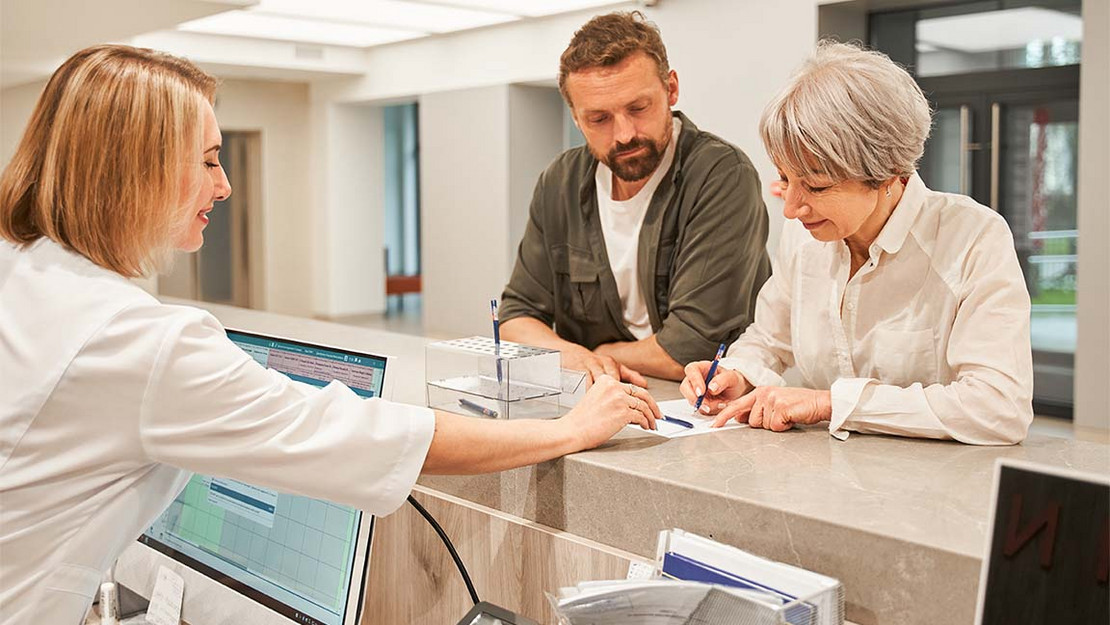 Eine ältere Frau und ein jüngerer Mann stehen am Empfangstresen einer medizinischen Einrichtung. 