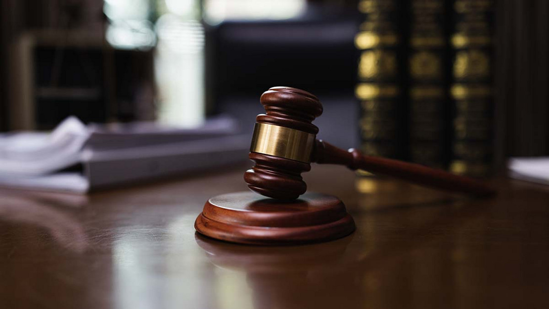 Ein Richterhammer liegt auf einem Pult, im Hintergrund Gesetzessammlungen. 