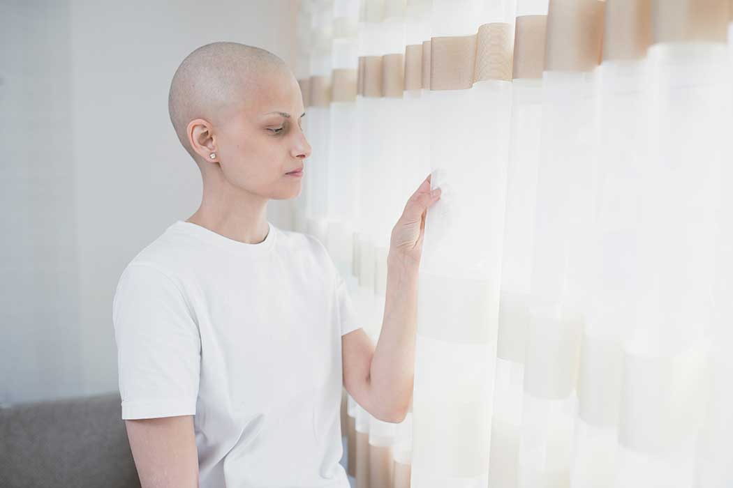 Frau mit Krebserkrankung steht an einem Fenster mit geschlossenen Vorhängen. 